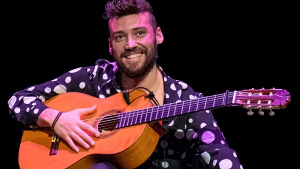A man with dark hair wearing a black shirt with white spots on it, sits smiling holding an acoustic guitar.