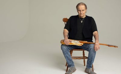 A man seated on a wooden chair wearing a black t-shirt, blue jeans and grey lace up boots holds an electric guitar over his lap. He sits in front of a grey background looking towards the camera.