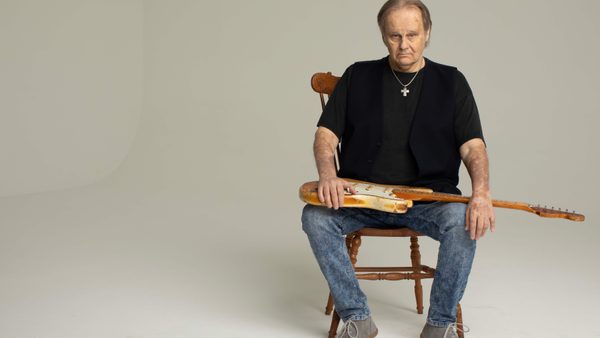 A man seated on a wooden chair wearing a black t-shirt, blue jeans and grey lace up boots holds an electric guitar over his lap. He sits in front of a grey background looking towards the camera.