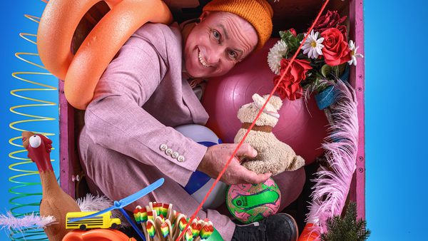 Person in a pink suit and orange beanie sitting in a cluttered space with colourful objects including a pool noodle, exercise ball, stuffed animal, flowers, and toys