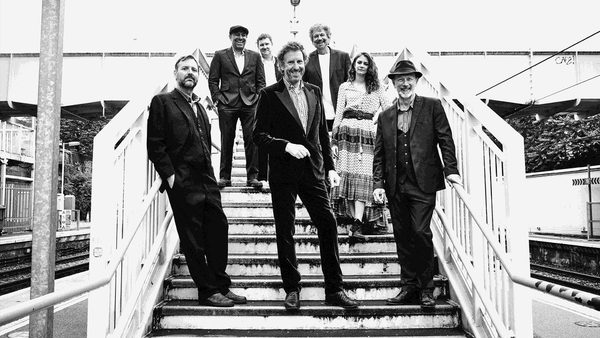 Group of seven people in formal attire standing on a staircase at a train station. Faces are obscured or blurred. Background includes train tracks, platforms, and an overpass bridge.