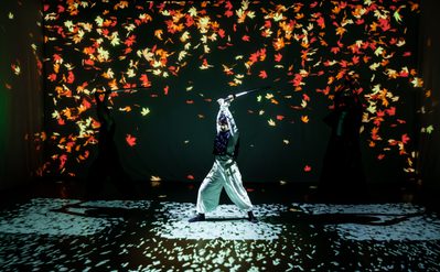 Performer in white trousers and dark top holding a raised sword on a stage with vibrant projections of falling red and orange leaves against a dark background.
