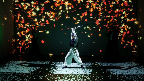 Performer in white trousers and dark top holding a raised sword on a stage with vibrant projections of falling red and orange leaves against a dark background.