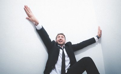 Seann Walsh wearing a black suit and tie leaning against a white wall with arms stretched wide, one arm raised upward and the other extended sideways.