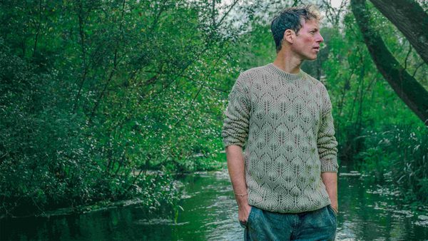 A person standing near a body of water in a lush green forest, wearing a patterned knitted sweater and blue jeans with hands in pockets.