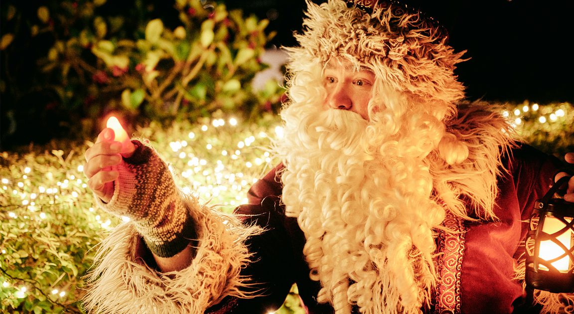 Person in a Santa Claus costume with a blurred face, holding a small lantern and a glowing object, surrounded by greenery and festive decorative lights.
