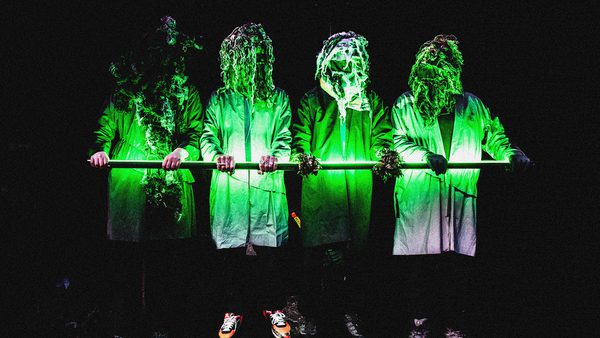 Four individuals in lab coats holding a horizontal bar, with their heads covered in foliage or plant material. The scene is illuminated with green light against a dark background, creating an eerie effect.