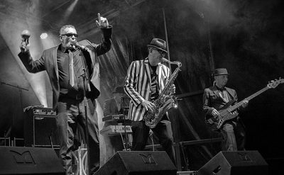 A black and white image showing three men - one is singing, one is playing a saxophone, and another is playing the bass guitar.