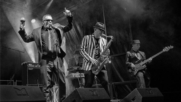 A black and white image showing three men - one is singing, one is playing a saxophone, and another is playing the bass guitar.