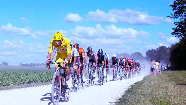 A group of cyclists racing on a gravel road under a bright blue sky with scattered clouds. The leading rider wears a yellow jersey and helmet, followed by others in colorful gear. Green fields and trees line the road, with spectators watching from the roadside as dust rises from the cyclists' wheels.