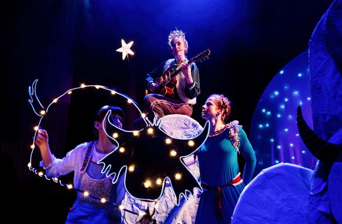 Three performers on a whimsical stage with blue lighting; one plays guitar on a platform, another holds a lit animal-head prop.