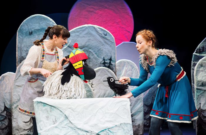Two performers in folk-style costumes holding bird puppets on a snowy mountain-themed stage with colorful lighting.