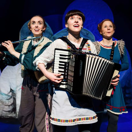 Three performers in colorful costumes playing flute, accordion, and singing on a winter-themed stage.