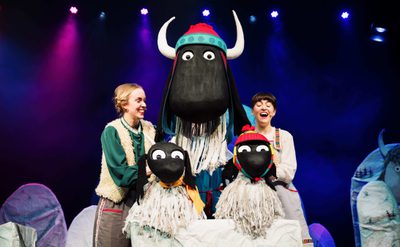 Two performers on stage interacting with large sheep puppets under colorful lighting.