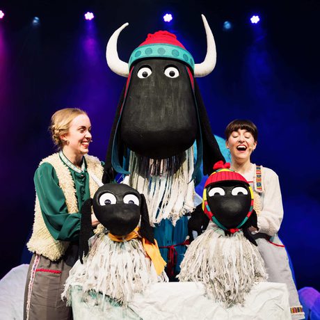 Two performers on stage interacting with large sheep puppets under colourful lighting.