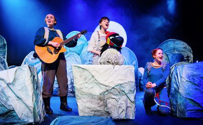 Three performers on a winter-themed stage with guitar, bird puppet, and icy props under blue lighting.
