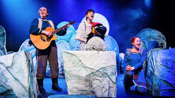 Three performers on a winter-themed stage with guitar, bird puppet, and icy props under blue lighting.