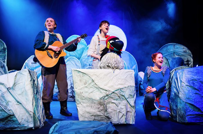 Three performers on a winter-themed stage with guitar, bird puppet, and icy props under blue lighting.