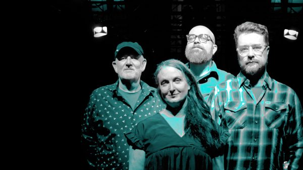 Four people with standing together; one wears a patterned shirt and cap, others wear a plain shirt, a dress, and a checkered shirt. Lights are visible in the background.