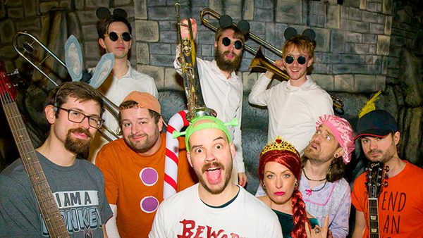 Group of people in costumes standing in front of a stone wall, with some holding musical instruments including a trombone and a guitar.