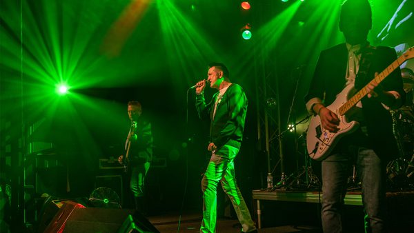 Three musicians performing on a stage illuminated with green lights; one singing into a microphone, another playing an electric guitar, and a third musician in the background, creating an energetic concert atmosphere.