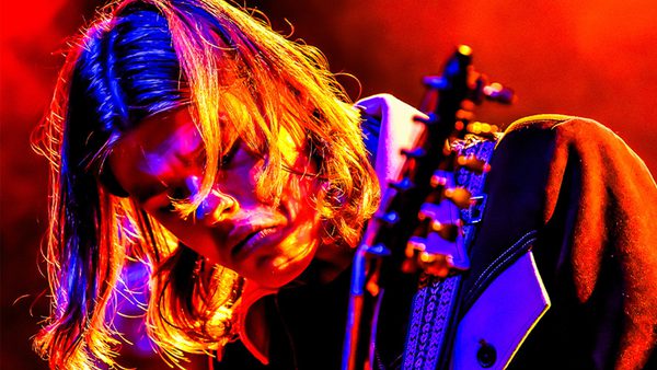 A long haired man playing a guitar with a red-lit background.