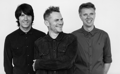 Three people standing side by side against a plain white background, wearing button-up shirts. The person in the centre has arms crossed, while the others stand with hands relaxed.
