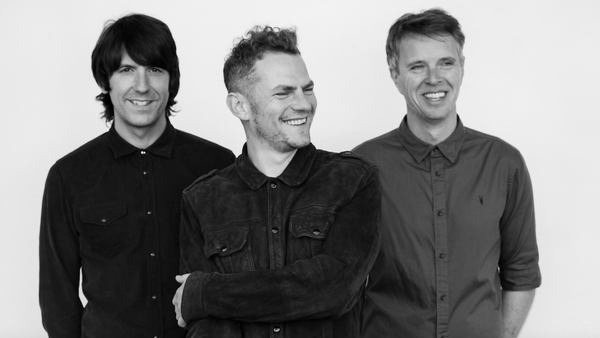 Three people standing side by side against a plain white background, wearing button-up shirts. The person in the centre has arms crossed, while the others stand with hands relaxed.