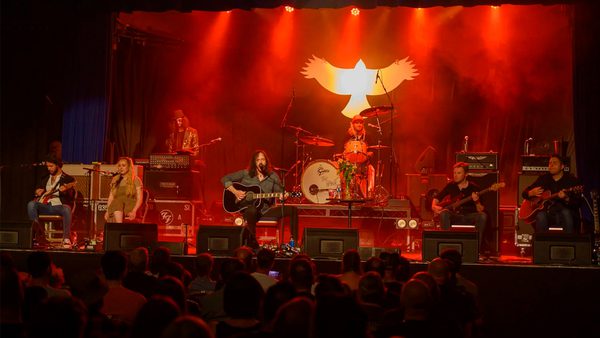 Live music performance on a stage lit with red lighting, featuring several musicians playing instruments; a large illuminated white bird symbol is in the background, with the audience visible in the foreground.