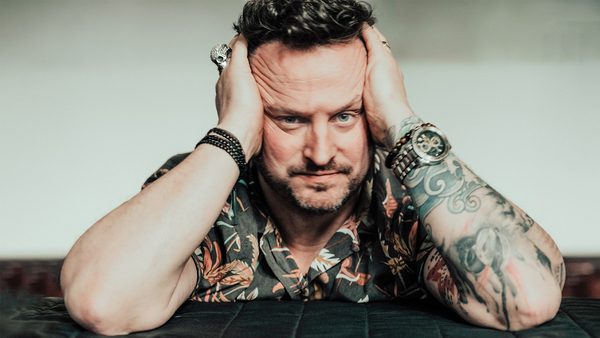 A person with tattooed arms wearing a floral shirt, bracelets, and rings, resting their head in their hands with elbows on a surface.