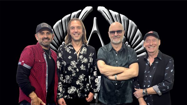 Four individuals in casual clothing standing in front of a black background with a large, stylized winged emblem featuring upward-curving wings meeting at the top center.