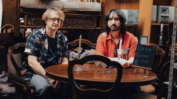 Two men sit at a round wooden table in what appears to be a second-hand furniture or thrift store, surrounded by assorted chairs, shelves, and stacked items. The man on the left has curly, light brown hair, wears glasses, and is dressed in a black shirt with a patterned open button-up. The man on the right has long dark hair, a beard, and is wearing a red short-sleeve shirt with a plaid pocket. On the table are a few wine glasses, candlesticks, and a small chalkboard sign that reads "CASH Payment ONLY." The background is cluttered with furniture and miscellaneous items, creating a cozy, vintage atmosphere.