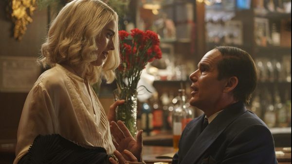 A woman with short blonde hair wearing a white blouse is standing talking to a seated man wearing a dark grey suit, she is holding a bunch of red flowers.