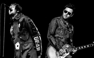 A black and white photo of two performers. One is singing in to a microphone and the second is playing an electric guitar.