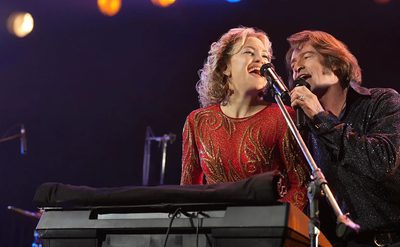 Two performers sing into the same microphone on stage. One wears a red glittery outfit and stands at a keyboard; the other wears a dark sparkly shirt. Colorful lights shine behind them.