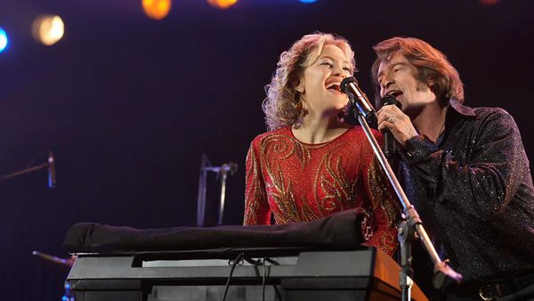 Two performers sing into the same microphone on stage. One wears a red glittery outfit and stands at a keyboard; the other wears a dark sparkly shirt. Colorful lights shine behind them.