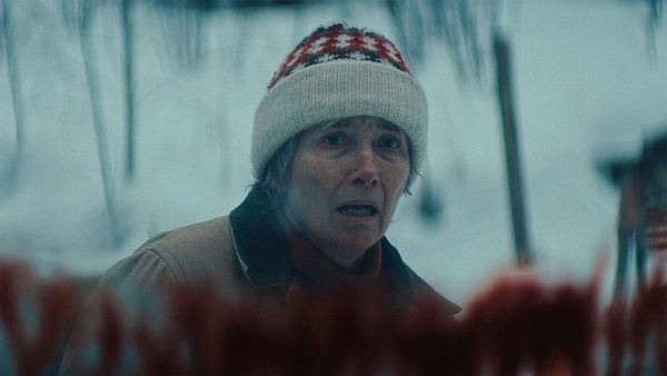 A person wearing a red and white patterned knitted hat stands outdoors in a snowy setting, looking intently off-camera against a blurred wintry background.