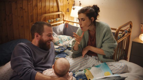 Three people in a warmly lit bedroom with a wooden crib. A baby is inside the crib, one adult is holding the baby, and another is gesturing while seated on the bed next to an open book.