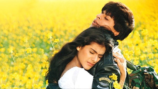 Two people embracing in a field of yellow flowers under a bright sky. One wears a white top with long dark hair, the other a black jacket with patches and short dark hair.