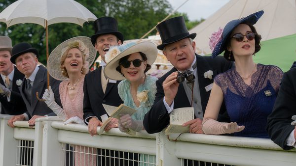 A group of people in formal attire stand behind a white fence. Some wear top hats and hold papers or programs, while one person holds an umbrella. The background features greenery and a tent-like structure.
