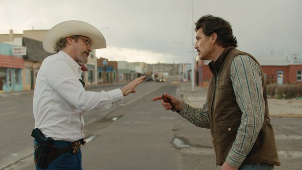 Two individuals standing on a street in a small town. One wears a white cowboy hat, white shirt, and jeans with a holster belt; the other wears a brown vest over a patterned shirt and jeans, pointing an object at the first person, who has a hand raised in a calming gesture.
