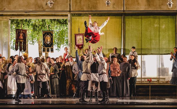 Large group of people in traditional clothing gathered in a festive setting, with several individuals lifting another person into the air; banners and decorations in the background suggest a cultural or historical celebration.