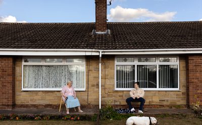 An older person sits on a chair with a cane. A younger person sits nearby with a white dog.