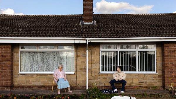 An older person sits on a chair with a cane. A younger person sits nearby with a white dog.