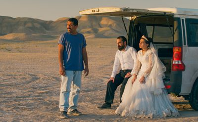 Three people are in a desert-like area with mountains in the background. One stands wearing a blue t-shirt and light trousers. Two others sit at the open back of a white van — one in a white wedding dress with a veil, the other in a white shirt and dark trousers. The lighting is warm, suggesting sunset or sunrise.