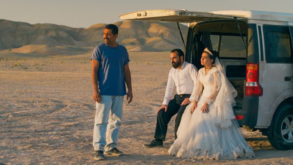 Three people are in a desert-like area with mountains in the background. One stands wearing a blue t-shirt and light trousers. Two others sit at the open back of a white van — one in a white wedding dress with a veil, the other in a white shirt and dark trousers. The lighting is warm, suggesting sunset or sunrise.