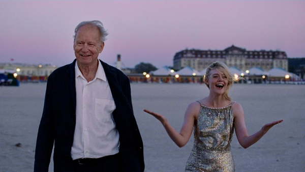 Two people walk along a beach at sunset, one in a dark jacket and one in a shiny dress.