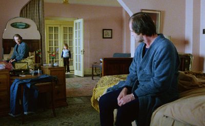 A man in a blue bathrobe sits on a bed in a pink bedroom. A child stands in the doorway wearing a blue and white sweater. The room has vintage furniture and soft daylight.