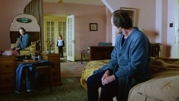 A man in a blue bathrobe sits on a bed in a pink bedroom. A child stands in the doorway wearing a blue and white sweater. The room has vintage furniture and soft daylight.