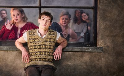 A person sitting on a wooden surface in front of a window, wearing a patterned knitted vest over a light shirt. Behind the window, several people are visible, with a wine bottle placed on the ledge.
